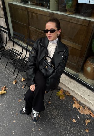 Chaqueta oversized de cuero negro con cremalleras, suéter blanco de punto, pantalones negros y zapatillas multicolores. Sentado en una silla al aire libre.