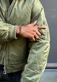 Giacca bomber verde oliva con tasca con zip sulla manica, polsini a costine e materiale in nylon lucido, caratterizzata da una chiusura frontale con zip e un design voluminoso.