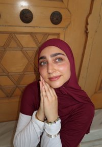 Burgundowa chusta owija twarz; pod nią nosi białą koszulę z długim rękawem. W tle widać drewnianą panelę z geometrycznym wzorem.