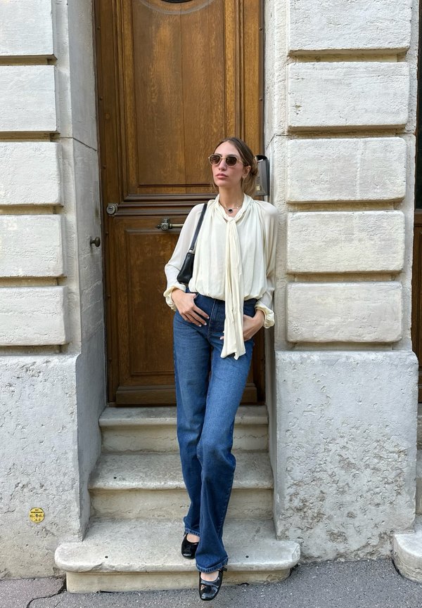 Mujer con gafas de sol, blusa blanca, vaqueros azules y zapatos negros que está de pie sobre unos escalones de piedra frente a una puerta de madera situada en una pared de piedra con textura.