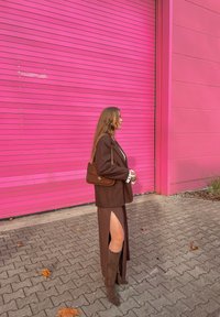 Veste marron portée sur une robe assortie avec une fente haute jusqu'à la cuisse, associée à des bottes marron montant jusqu'au genou et un sac à bandoulière en suède, devant un mur rose.