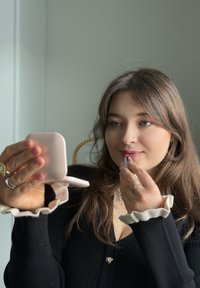 Jeune femme appliquant un rouge à lèvres rose à l'aide d'un miroir de poche, portant un haut noir avec des poignets blancs à volants et des bagues aux doigts.
