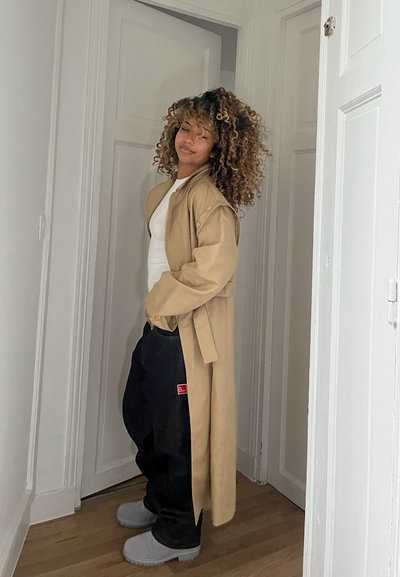 Abrigo largo beige de corte suelto, top blanco, pantalones negros anchos con un parche rojo y botas grises con suela texturizada. Suelo de madera, paredes blancas.