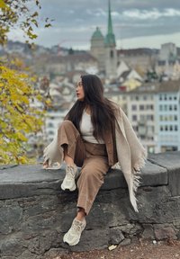 Un mannequin est assis sur un mur de pierre, portant un ensemble en velours côtelé marron, un haut crème, et des baskets blanches, enveloppé dans un châle beige clair à franges.