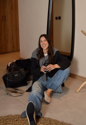 Mujer joven sentada en el suelo con una taza de café, vestida con vaqueros y un blazer negro, cerca de un bolso negro y zapatos beige, sonriendo a la cámara.