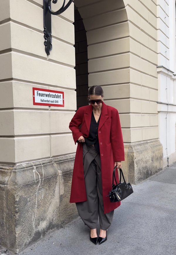 Mujer con abrigo rojo, blusa negra, pantalones grises y tacones negros que sostiene un bolso negro, de pie junto a un edificio de piedra beige bajo un arco.
