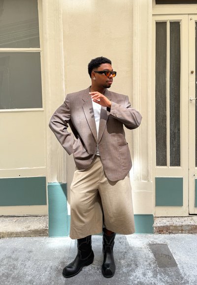 Blazer de cuadros marrón de corte oversize, camisa blanca, culottes beige y botas negras hasta la rodilla. Unas gafas de sol y pendientes de botón completan el look.