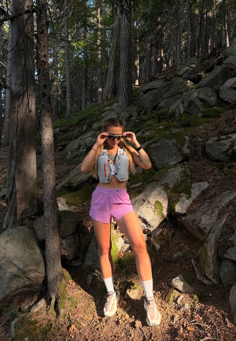 Femme portant un short rose et des baskets blanches ajustant ses lunettes de soleil tout en se tenant sur un sol forestier rocheux avec des arbres et des motifs de lumière.