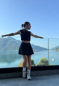Fekete öltözetbe öltözött nő karjait nyújtja egy üveg erkélyen, amely a nyugodt tóra és a hegyekre néz a tiszta kék ég alatt.