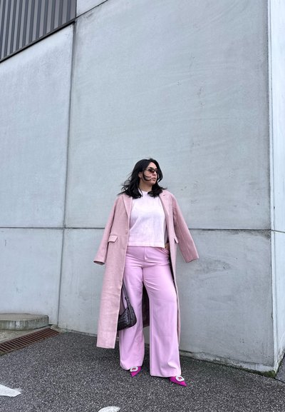 Mujer con abrigo largo rosa, top blanco y pantalones anchos rosa, de pie contra una pared de hormigón, con gafas de sol y sosteniendo un bolso negro.