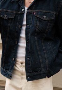 Giacca di jeans scura con cuciture in rame, due tasche sul petto e chiusura con bottoni, indossata sopra una maglietta bianca e pantaloni beige.