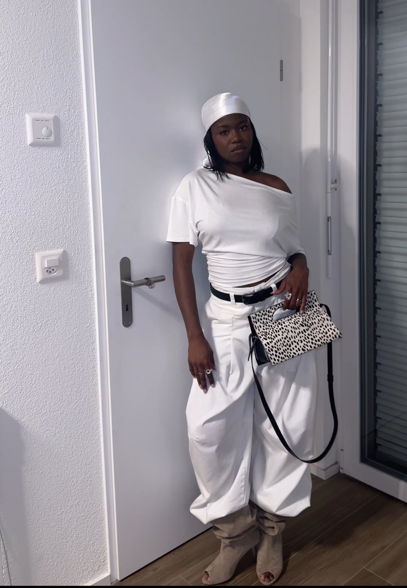 Kobieta ubrana w biały top odkrywający ramiona, białe spodnie, beżowe botki, biały chustę na głowie, trzyma czarną i białą torebkę w cętki, opierając się o białe drzwi.