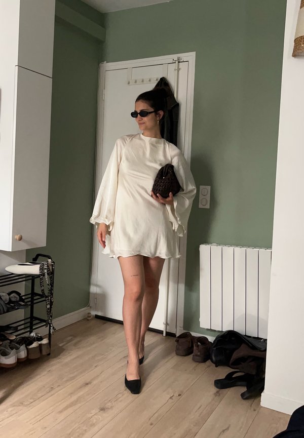 Žena v bielych šatách s dlhým rukávom a čiernymi slnečnými okuliarmi a čiernymi podpätkami stojí v interiéri a drží malú tmavú kabelku pri zelenej stene.