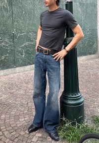 Person iført en mørkegrå tætsiddende T-shirt, brede blå jeans, sorte sko og bælte, der står med hænderne på hofterne og læner sig op ad en grøn gadelygte.