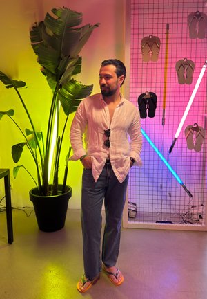 Hombre vestido con camisa blanca, vaqueros y chanclas está junto a una planta grande y una pared de exhibición con chanclas colgadas y luces de neón.