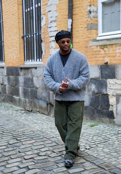 Hombre con boina negra, gafas de sol redondas, cárdigan gris y pantalones verdes caminando por una calle empedrada junto a una pared de ladrillos amarillos con ventanas con rejas.