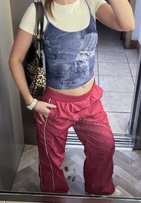 Γυναίκα που φοράει τσάντα με λεοπάρ μοτίβο, λευκό μπλουζάκι σε στρώσεις και κοντό τζιν τοπ, κόκκινο αθλητικό παντελόνι, βραχιόλι και ρολόι, στέκεται σε ένα δωμάτιο με πλακάκια.