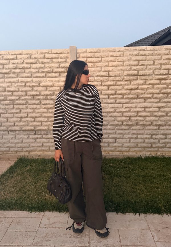 Femme portant des lunettes de soleil noires, une chemise à manches longues rayée, un pantalon large et foncé, tenant un sac à main noir, debout près d'un mur en pierre et d'une parcelle d'herbe.