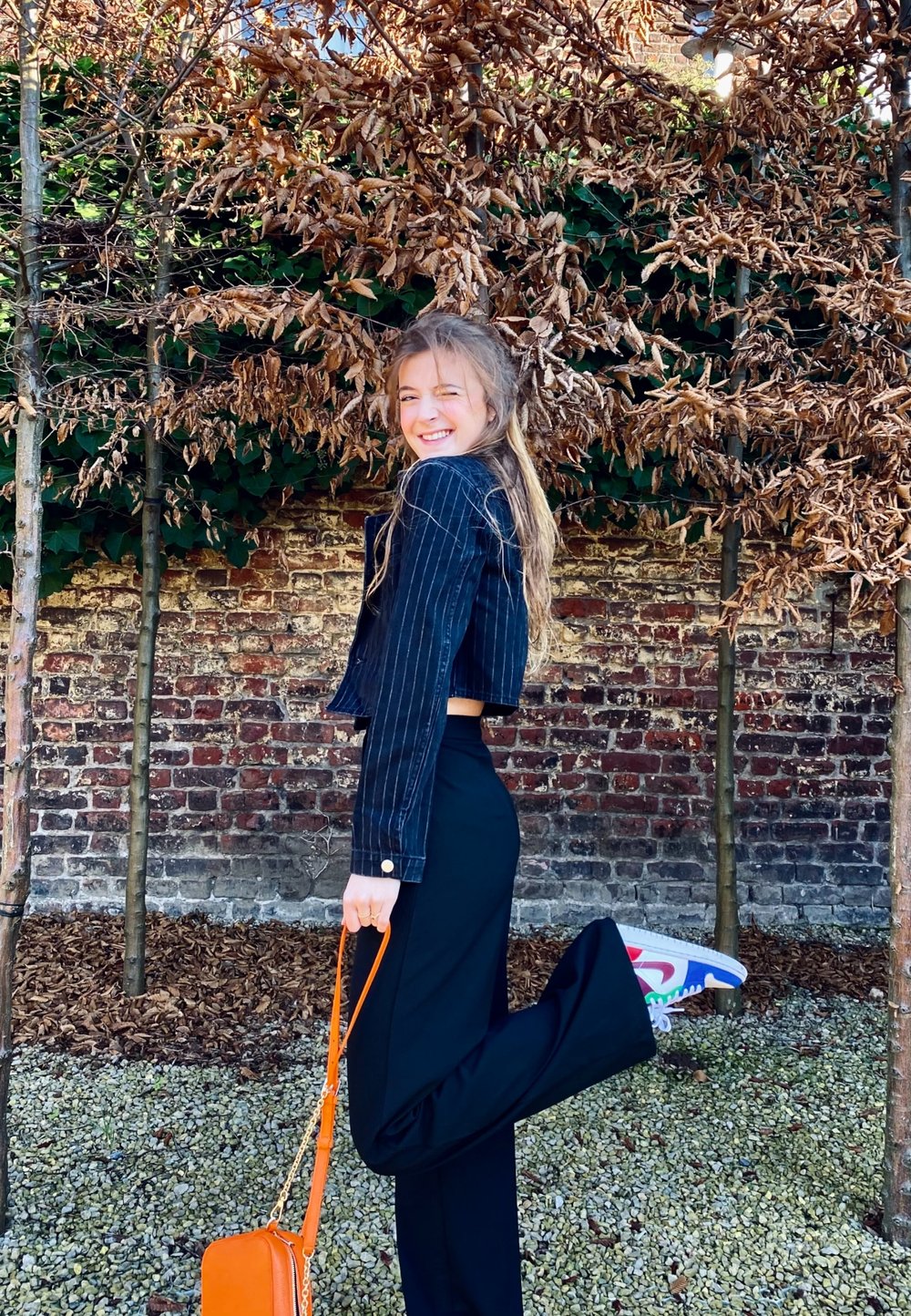 Sort jakkesæt med prikker, sorte bukser, farverige sneakers og en orange håndtaske. Baggrunden har bare træer og en murstensvæg.