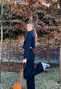 Sort jakkesæt med prikker, sorte bukser, farverige sneakers og en orange håndtaske. Baggrunden har bare træer og en murstensvæg.