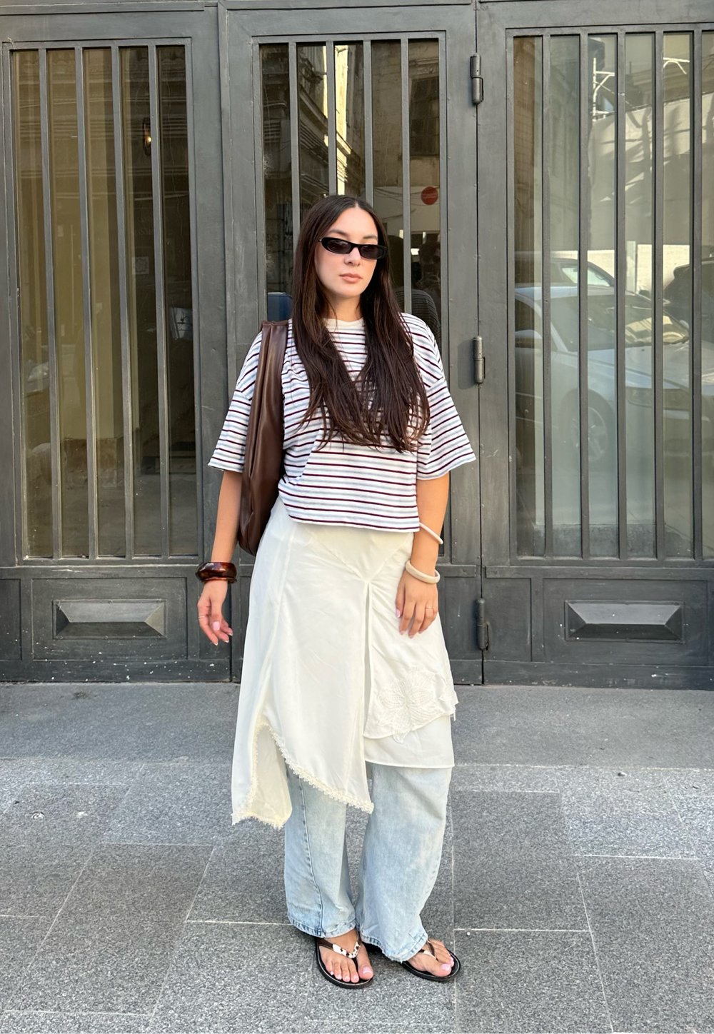 Stribet kortærmet top, cremet lagdelt nederdel, lys blå vide jeans, brun håndtaske og sorte sandaler med tåstykke. Baggrunden har rutenetmønstrede døre.