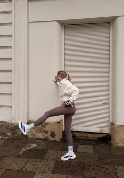 Mujer con ropa deportiva estirando la pierna contra una pared en una acera urbana mojada y con baldosas.