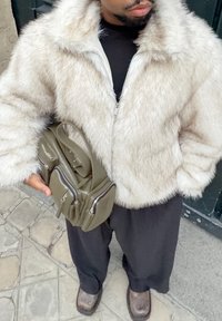 Veste en fausse fourrure beige clair, fermeture éclair, manches larges. Sac à main vert olive avec plusieurs fermetures éclair et une poignée courte, accompagné d'un pantalon foncé.