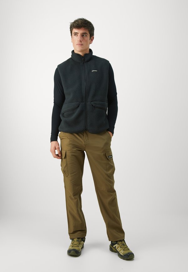 Black fleece vest with a high collar and two front pockets, worn over a long-sleeve black shirt, teamed with olive cargo pants and hiking shoes.