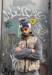Homme aux bras croisés portant des lunettes de soleil orange, un bonnet noir avec un imprimé blanc, un gilet Adidas camouflage et un sweat à capuche gris, debout devant une porte en métal couverte de graffitis.