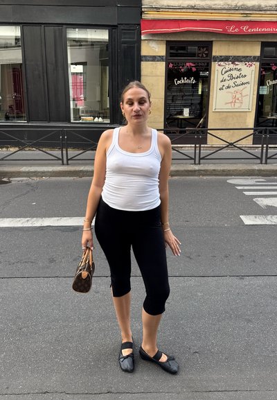 Top sin mangas blanco y leggings capri negros. Sosteniendo un bolso marrón con estampado, lleva bailarinas negras con una tira. Entorno urbano.
