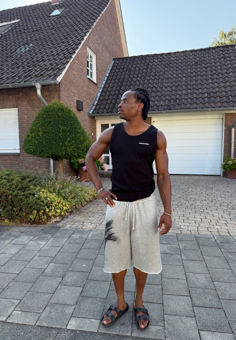 Homme en débardeur noir, short gris et sandales, se tient avec une main sur la hanche devant une maison en brique avec toit en tuiles et garage.