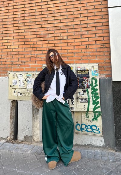 Chaqueta negra de gran tamaño, camisa blanca, corbata negra, pantalones de satén verde, botas marrones, gafas de sol, de pie contra una pared de ladrillo con graffiti.