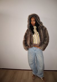 Veste en fausse fourrure marron avec capuche, associée à un haut à motifs jaune et bleu et un jean large bleu clair. Le sol est en bois clair.