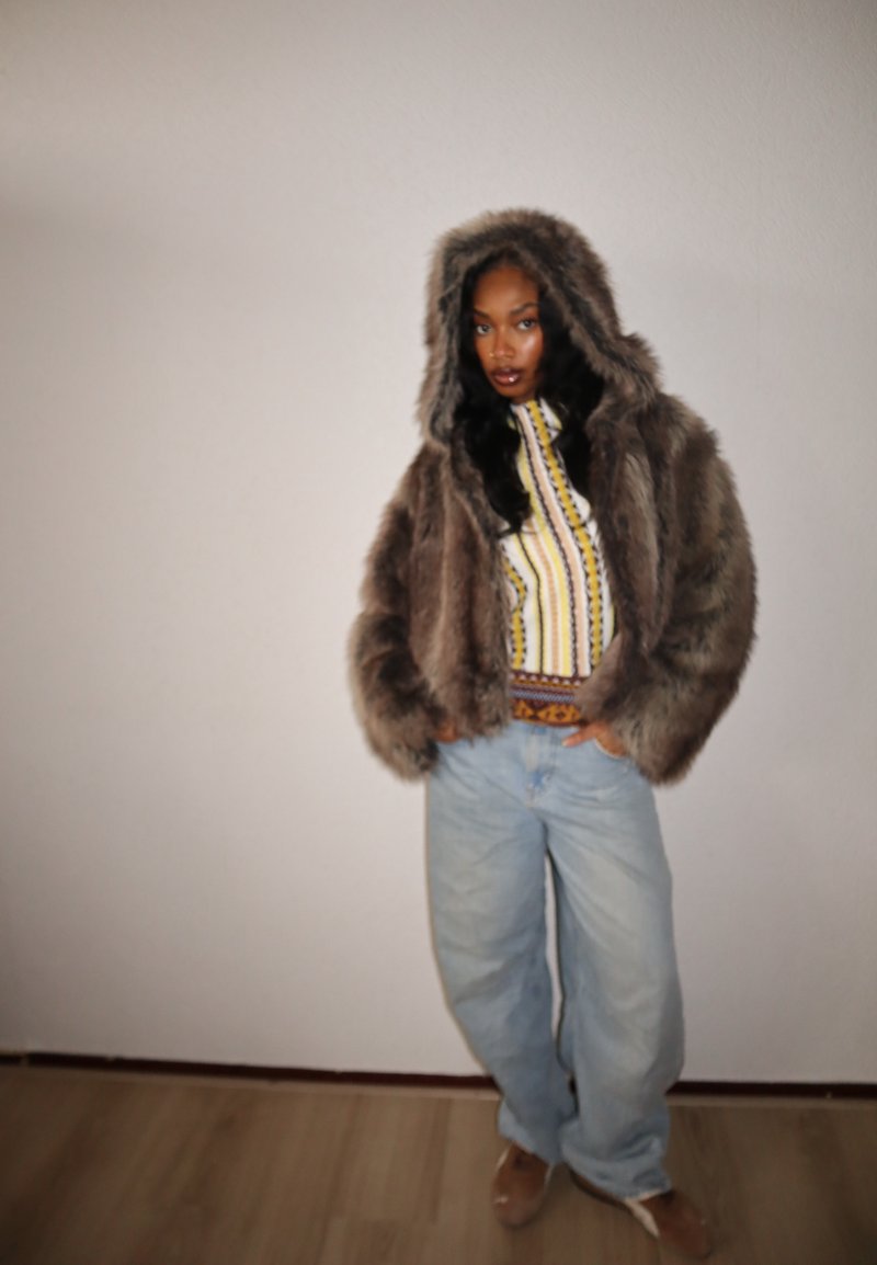 Giacca marrone in pelliccia sintetica con cappuccio, abbinata a una maglia gialla e blu con motivo e a jeans larghi color azzurro chiaro. Il pavimento è in legno chiaro.