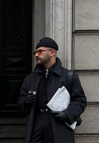 Άνδρας με μαύρο παλτό και γάντια, κρατώντας μια εφημερίδα και ένα φλυτζάνι καφέ, στέκεται δίπλα σε έναν υφασμάτινο γκρι τοίχο και μαύρη πόρτα.