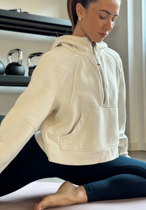 Sudadera corta beige con media cremallera y bolsillos laterales, combinada con leggings negros, colocada sobre una alfombra clara en un interior luminoso.