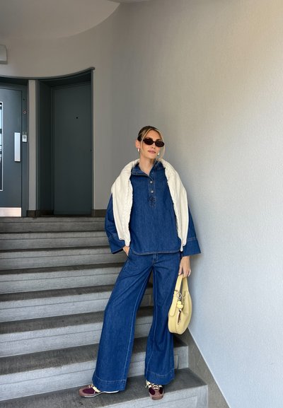 Conjunto de denim con una camisa azul oversized y pantalones holgados, superpuestos con un suéter crema. Los accesorios incluyen gafas de sol oscuras y un bolso amarillo.