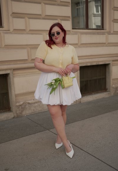 Cárdigan amarillo de manga corta con botones, falda semi-bolita blanca, tacones blancos de punta y bolso amarillo con tulipanes. Texturas suaves.