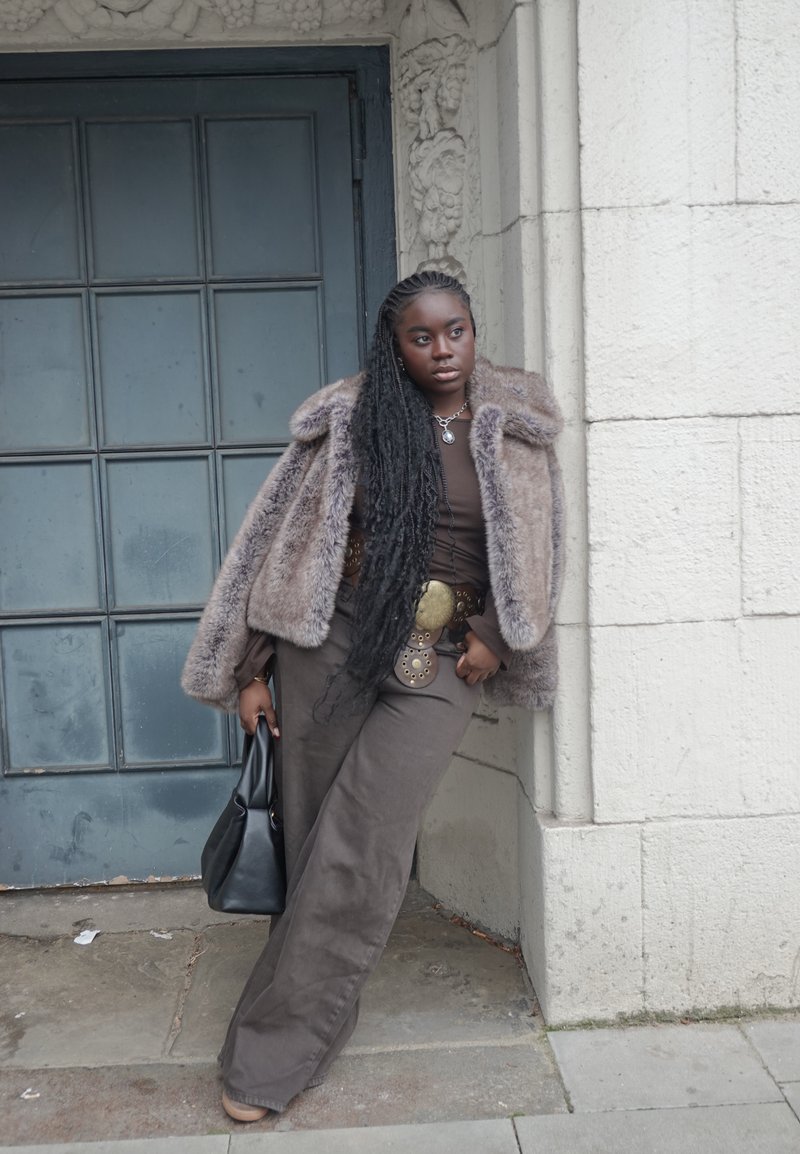 Manteau en fausse fourrure marron avec un large col, associé à un haut marron ajusté et un pantalon ample avec une ceinture décorative. Sac cabas noir.