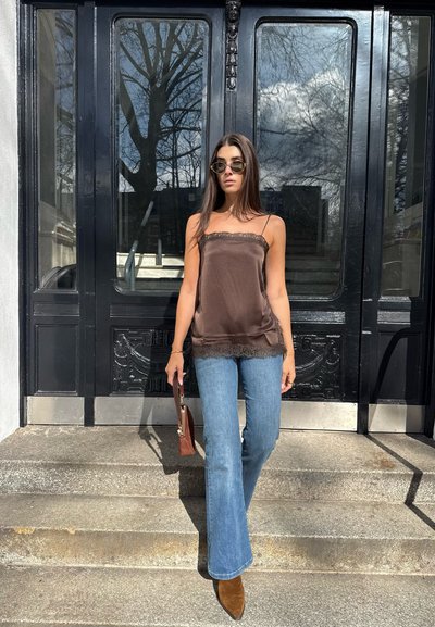 Mujer con cabello largo y oscuro, lleva gafas de sol, top marrón con encaje, vaqueros acampanados azules y botas marrones, de pie sobre escalones de piedra frente a puertas negras.