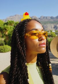 Óculos de sol amarelos com lentes tingidas, usados por uma pessoa com cabelo longo e torcido adornado com um acessório de cabelo em forma de pato de borracha amarelo. Fundo brilhante.