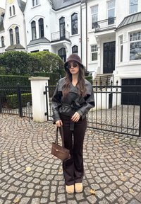 Femme portant un chapeau bob marron, une veste en cuir noire, un pantalon marron, des chaussures beige et des lunettes de soleil, tenant un sac à main marron dans une rue pavée.