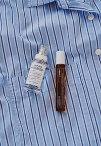 Tydlig glasflaska med spraymunstycke märkt "REPLICA" bredvid ett brunt rör märkt "KORRES," placerad på en blårandig skjorta.