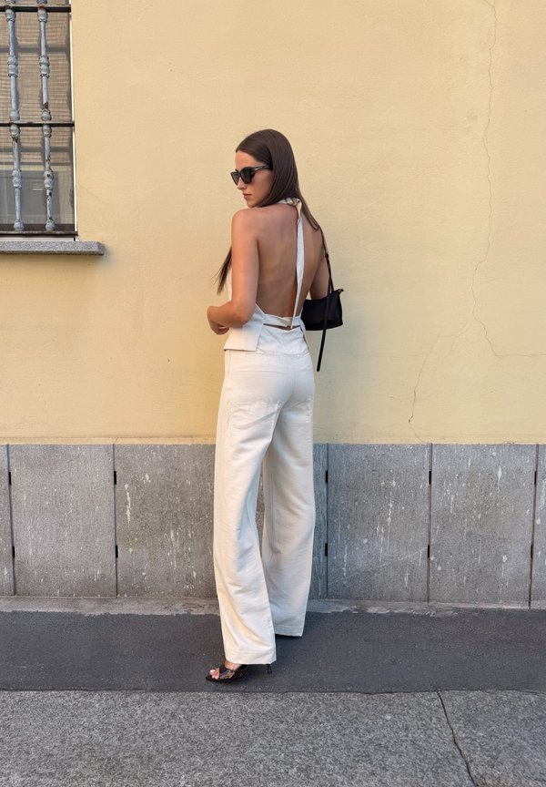 Femme portant un haut dos nu blanc à col halter et un pantalon blanc à jambes larges, debout contre un mur jaune avec une fenêtre grillagée.