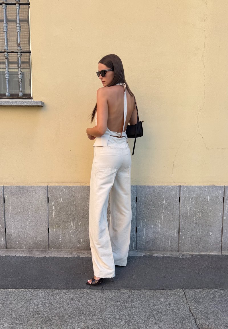 Donna che indossa un top bianco senza schienale con scollo all'americana e pantaloni bianchi a gamba larga, in piedi contro un muro giallo con una finestra con inferriata.