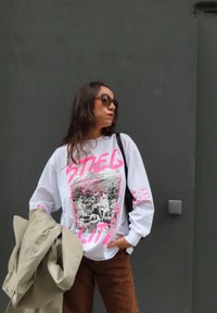 Bílá dlouhá tričko s long sleeves, na kterém se nachází černobílá grafika s růžovými akcenty. Hnědé kalhoty a béžová bunda přehozená přes ruku.