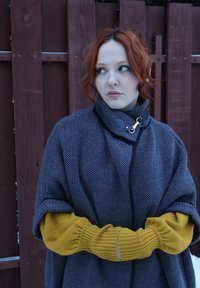 Femme aux cheveux roux et à la peau pâle portant une cape grise texturée et des manches jaune moutarde, debout les bras croisés contre une clôture en bois.