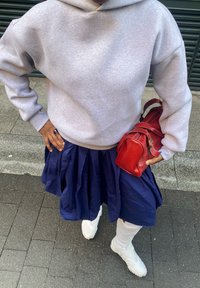 Pelēks kapucjakas džemperis, tumši zils svārks ar krokām un balti augstie sporta apavi. Rokā tur mazu sarkanu rokassomu ar zelta akcentiem. Redzama teksturēta auduma virsma.