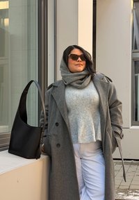 Manteau en laine gris avec un pull court gris clair et un pantalon blanc. Sac à bandoulière en cuir noir, le modèle porte des lunettes de soleil noires et une écharpe grise.