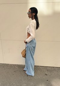 Top blanco de manga larga, jeans sueltos de color azul claro, gafas de sol de montura transparente, sosteniendo un bolso de mano marrón. Texturas suaves, ajuste casual, colores claros.